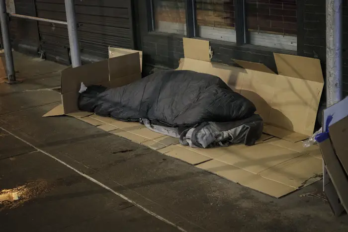 A person covered by a black blanket sleeps on some cardboard on a city sidewalk.